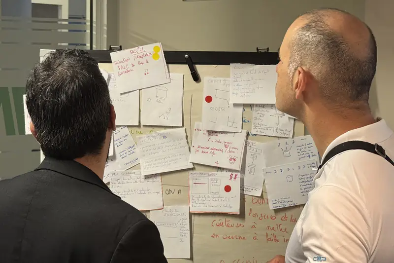 Deux participants observent un mur de croquis et de pistes, marqués de gommettes de vote, pendant un sprint de design