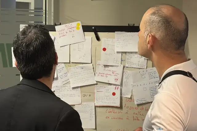 Deux participants observent un mur de croquis et de pistes, marqués de gommettes de vote, pendant un sprint de design